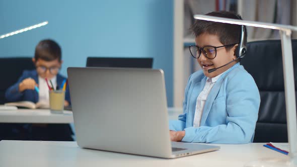 Little Confident Businessman Boy in Office Wearing Headset Talking Online on Laptop Sitting at Desk