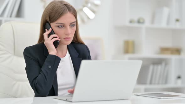 Serious Businesswoman with Laptop Talking on Smartphone in Office