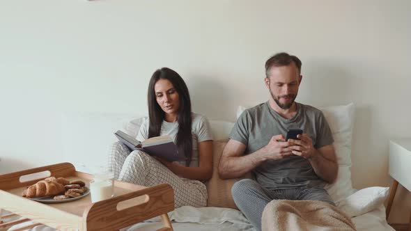 Couple in bed with breakfast tray, reading book, using smartphone