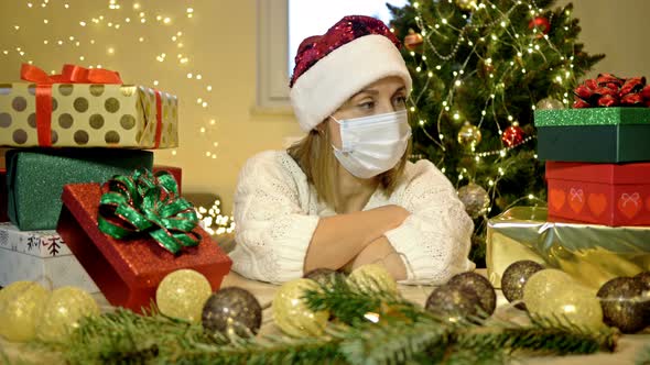 Sad Woman in a Santa Hat and a Protective Mask Sits Alone Against the Background of a Christmas Tree