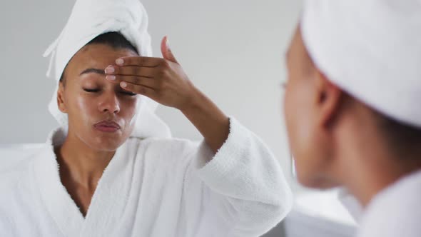 African american woman in bathrobe applying face cream looking in the mirror at bathroom