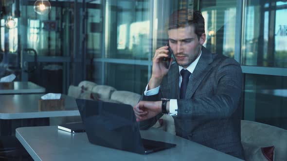 Young Businessman in a Suit Is Sitting on the Terrace of a Cafe and Working on a Laptop, Talking on