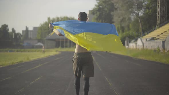 Confident Ukrainian Athlete Walking with National Flag Along Stadium and Turning To Camera. Portrait