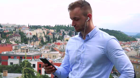 Young Handsome Business Man Listens Music with Earphone on Smartphone - City in Background