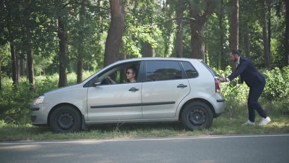 Young Caucasian Woman Sitting on Driver's Seat As Man Pushing Vehicle Outdoors on Sunny Day