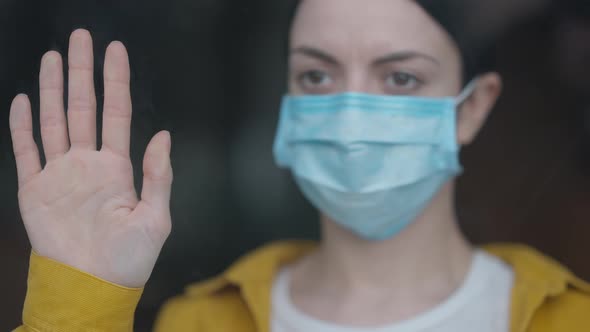 Closeup Female Hand on Glass with Blurred Sad Woman on Coronavirus Face Mask at Background