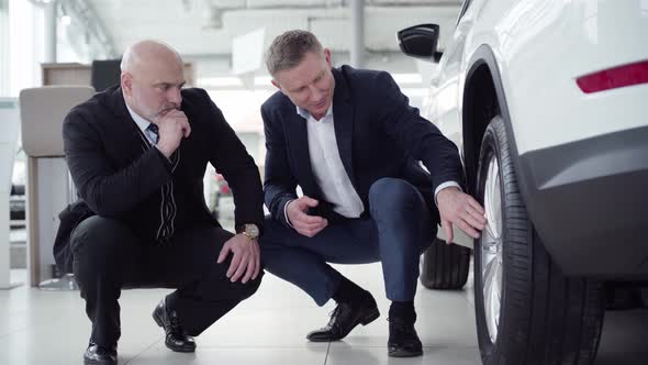 Serious Senior Caucasian Man Sitting on Hunkers with Car Dealer Next To New Vehicle