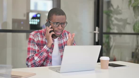 Angry African Man Talking on Smartphone While Using Laptop