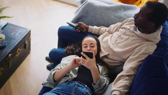 Overhead Shot Of Relaxed Young Couple At Home On Sofa Watching TV And Checking Mobile Phone