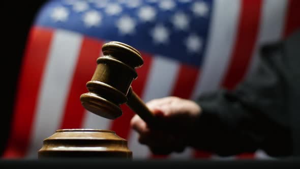 Judge Hammering With Wooden Gavel in Hand Against Waving American Flag in United States Court