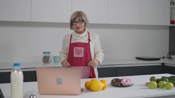 Front View Portrait of Confident Satisfied Female Retiree Putting on Apron Standing in Kitchen at