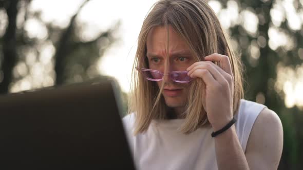 Portrait of Embarrassed Caucasian Young Man Putting Down Sunglasses Looking at Laptop Screen