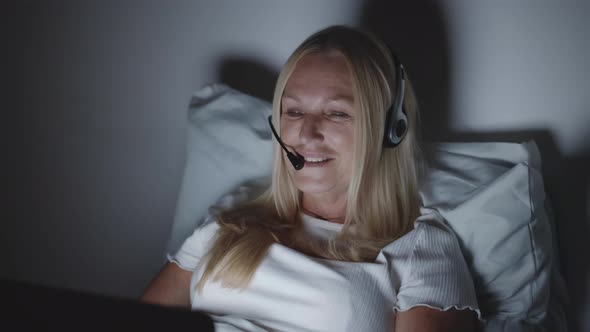 Mature Woman in Headset Having Video Call on Laptop Lying in Bed Late at Night