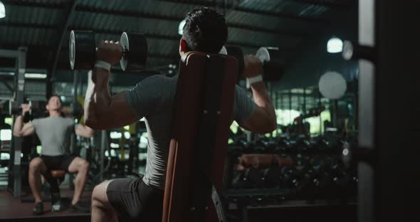 Black view of muscular man guy lifting two dumbbell during workout while sitting on bench