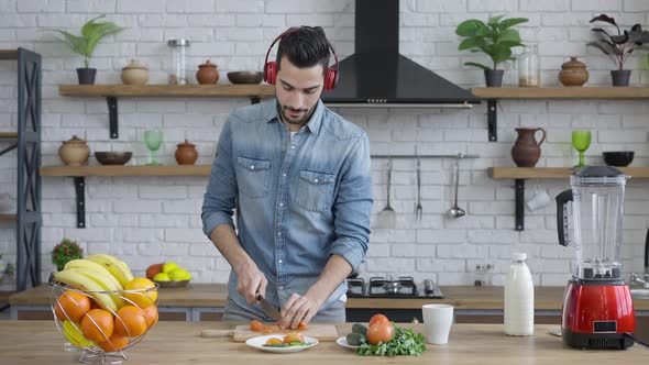 Handsome Joyful Middle Eastern Man in Headphone Cutting Tomato for Healthful Vegan Salad and Dancing