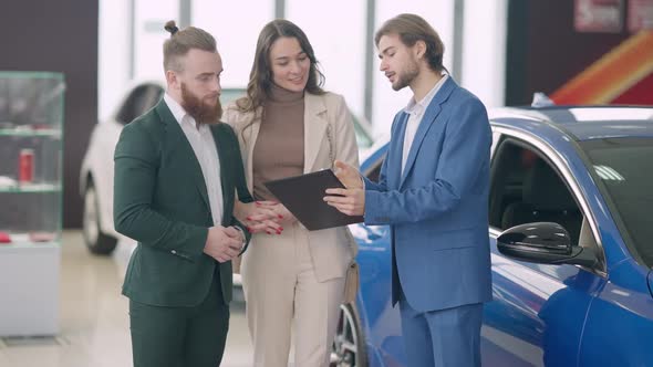 Confident Car Dealer and Smiling Young Wealthy Couple Standing in Dealership Talking