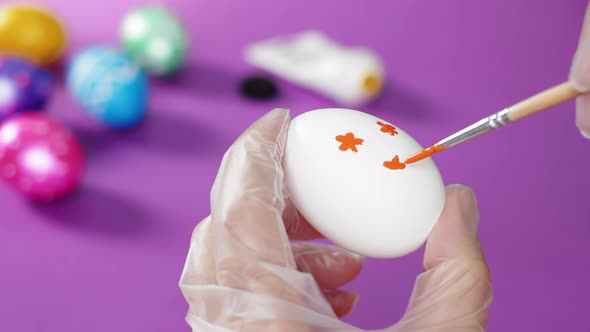 A man's hand, wearing a transparent glove, draws red flowers on a white Easter egg