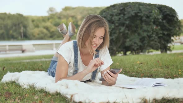 A Young Girl Lies on a White Plaid in the Park and Dials a Message on the Phone