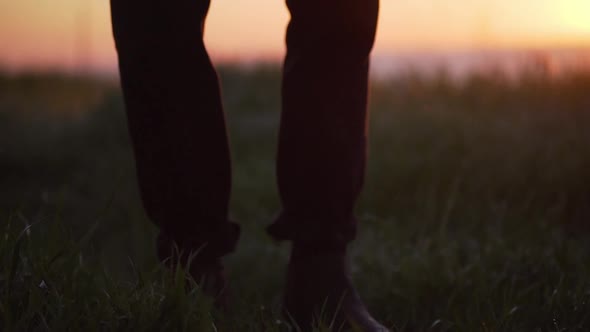 Panoramic Shot of Mens Legs in Boots and Trousers Standing in Grass with Sea Sunset in Background