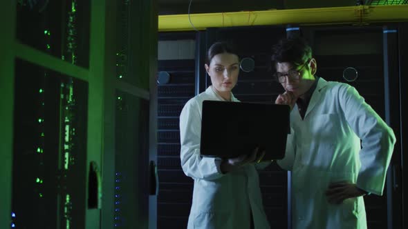 Diverse female and male it technicians in lab coats using laptop checking computer server