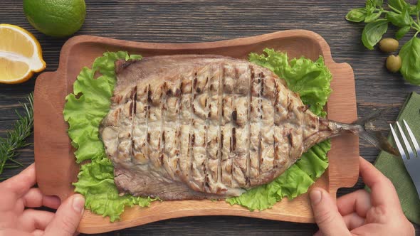 Hands are Putting Plate with Grilled White Mackerel Fish on a Dark Wooden Table