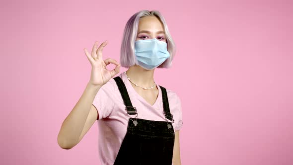 Cute Woman with Violet Dyed Hairstyle and Medical Mask Showing Okay Sign Over Pink Background