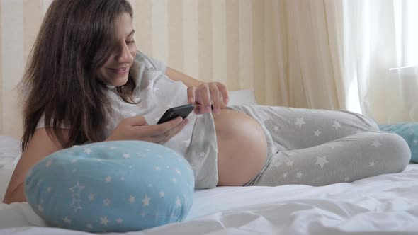 Pregnant Caucasian Woman Lies In Bed Uses A Smartphone And Strokes Her Stomach