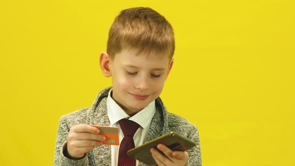 A cheerful Caucasian boy of 6-7 years old makes online payments using a bank card and a smartphone