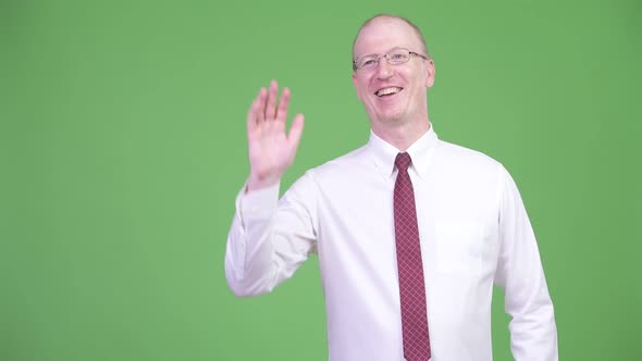 Mature Bald Businessman Smiling While Waving Hand Against Green Background