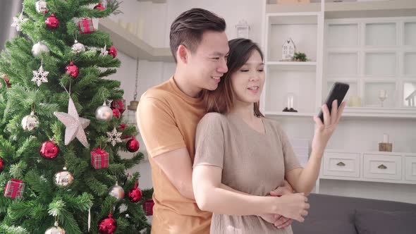 Asian couple using smartphone selfie with christmas tree decorate their living room in Christmas.