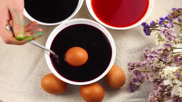 Caucasian Child Paints an Easter Egg with a Metal Spoon and Dips It Into a White Bowl with Red Dye