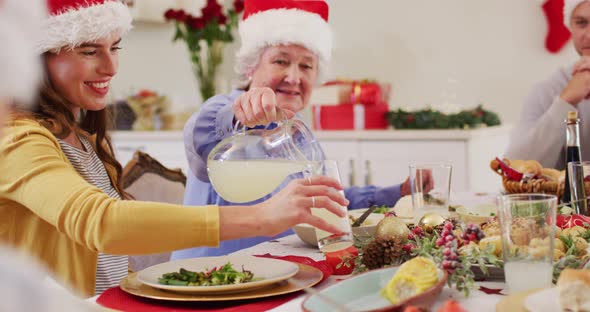 Caucasian senior woman in santa hat pouring drink in glass of woman while sitting on dining table an