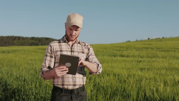 The Farmer is Standing Near the Field and Working with a Tablet