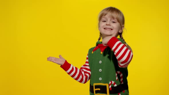 Kid Girl in Christmas Elf Santa Helper Costume Points Fingers at Blank Space Shows Advertising Area