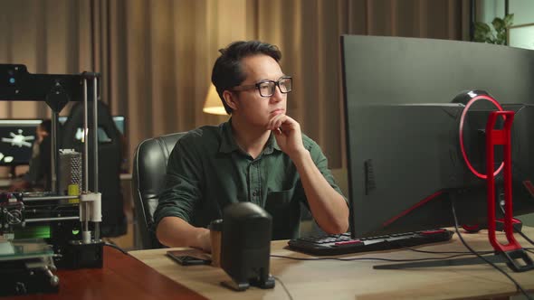 Asian Man Thinking While Works On Personal Computer And 3D Printer In Home Office