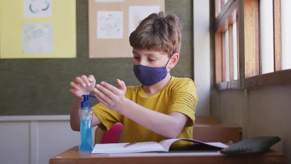 Boy wearing face mask sanitizing his hands while sitting on his desk at school