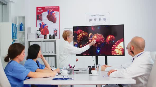 Senior Doctor Pointing on Monitor in Hospital Office Explaining Virus Development