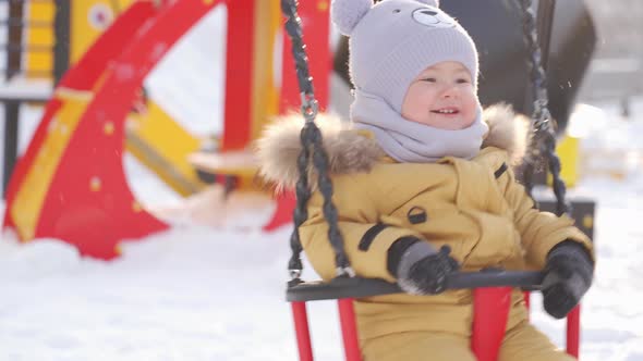A newborn baby in yellow winter clothes swings on a swing and laughs. Winter playground