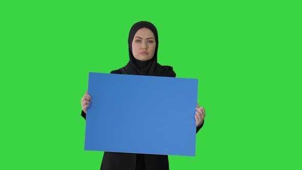 Serious Arab Woman in Hijab Holding Blank Blue Poster and Looking at Camera on a Green Screen