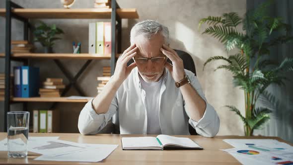 Middleaged Man Working at Home Office Looking at Notepad Being Puzzled and Massaging Temples Taking