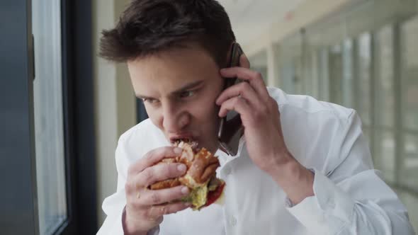 Young Man Eats Burger and Talks on Smartphone at the Window in Office Center