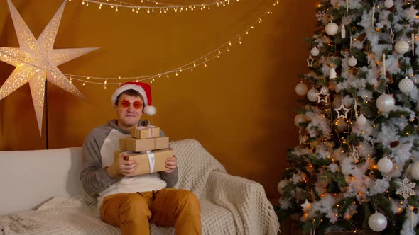 Man in Santa Hat and Bright Glasses Sitting with Wrapped Gift Boxes on Sofa Near Christmas Tree