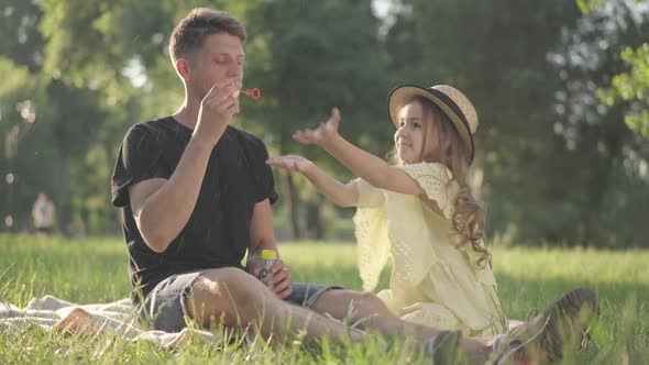 Joyful Caucasian Daughter Catching Soap Bubbles As Father Blowing Soap Sud Sitting on Green Spring