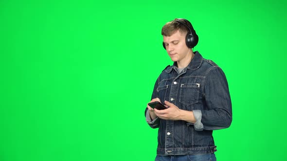 Man in Big Headphones Goes and Texting with Smartphone on Green Screen at Studio