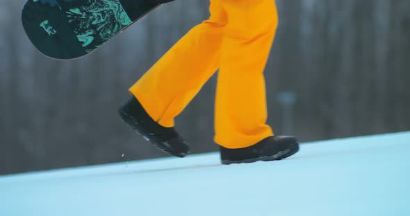 Snowboarder Rises Up the Track, Holds a Snowboard in His Hands. Snowboarder Legs Rise Uphill on a