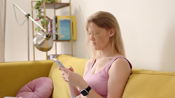 Blonde Sits on Yellow Sofa in Pink Top and Use Phone Typing Text