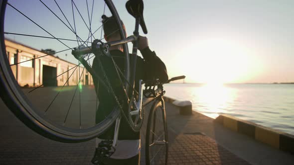 Stylish Handsome Man Walking with Bicycle on His Shoulder During Sunset or Sunrise with Sea Port on