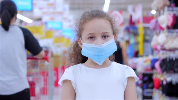 Cute Little Girl From an Epidemic of Coronaviruses or Viruses Looks at the Camera Amid Masked People