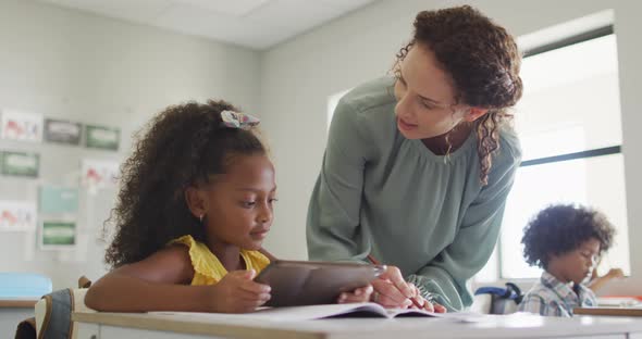 Video of happy caucasian female teacher explaining lesson to african american girl holding tablet