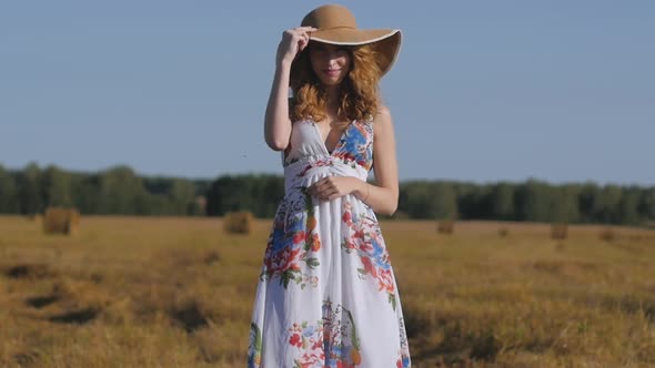 Redhead Girl in a White Dress and Hat on the Background of a Field and Haystacks